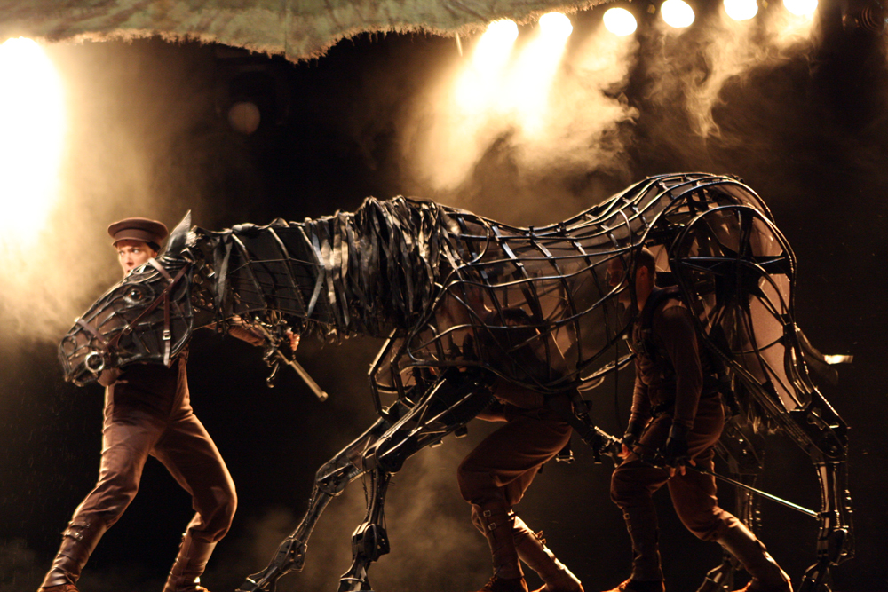 Side view of a skeletal War Horse puppet animated by three performers crossing a lit stage