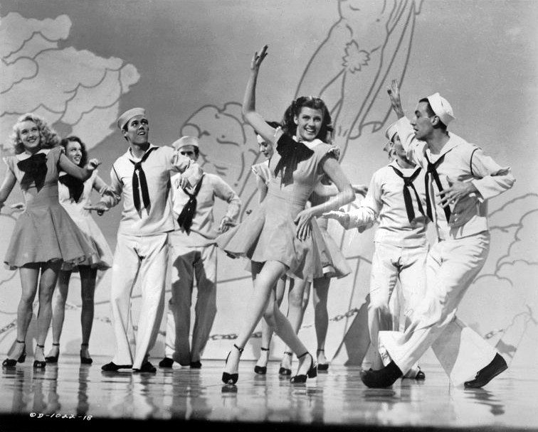 Rita Hayworth dancing with a sailor while Jack Cole stands at far right in a 1945 film still.