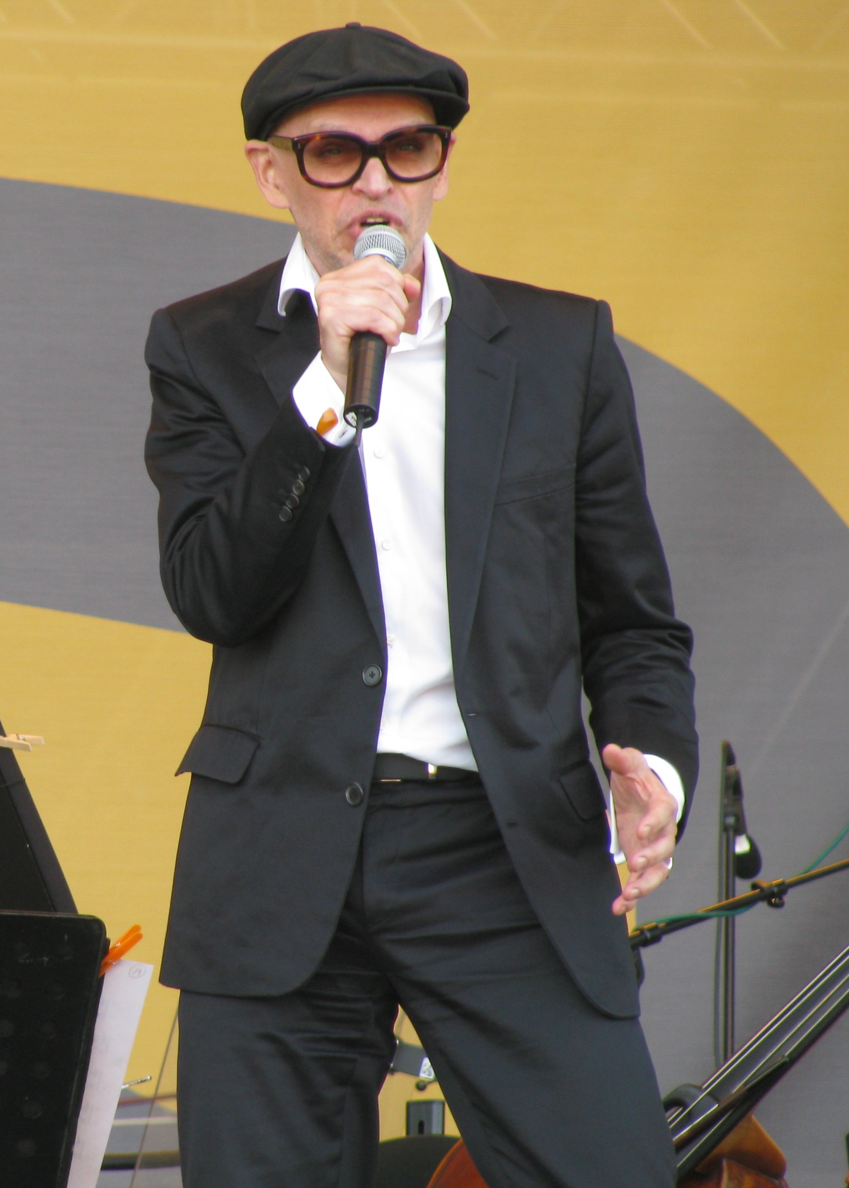 Jorma Uotinen standing on an outdoor stage in a black suit and cap, speaking or singing into a microphone in Pori in 2008.