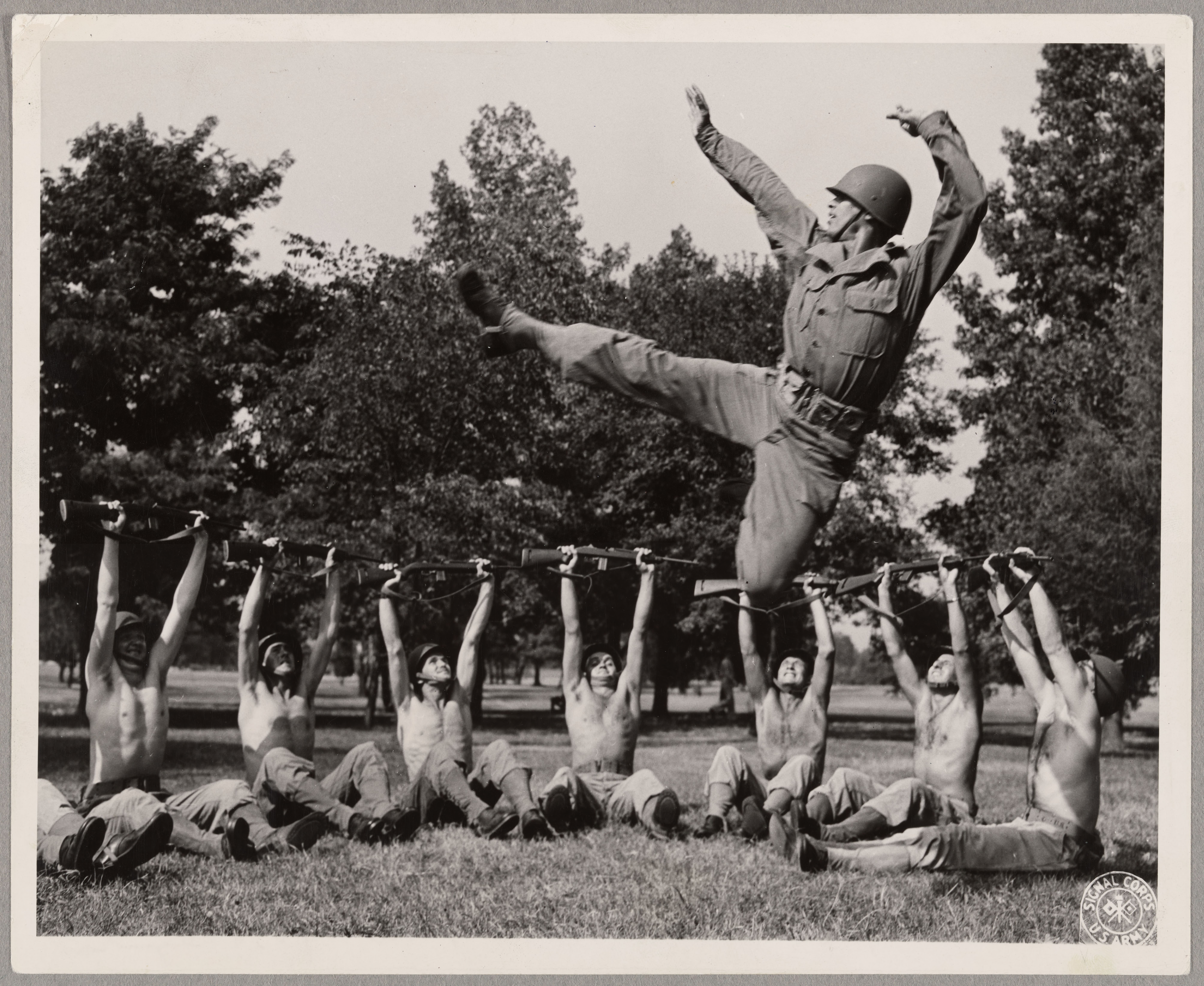 Black-and-white outdoor photograph of José Limón leaping in uniform above soldiers rehearsing with rifles in 1943.
