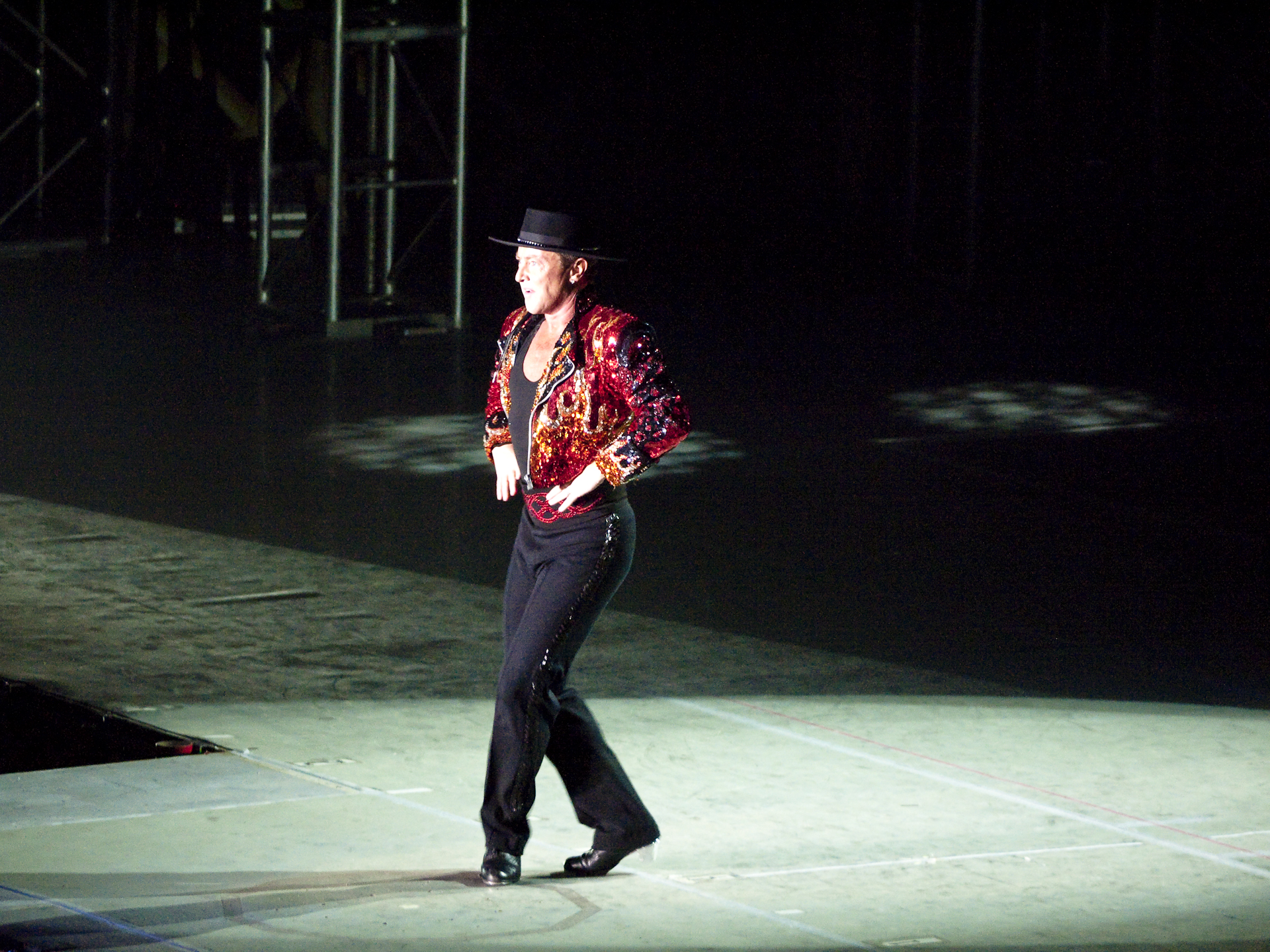 Michael Flatley dancing onstage in a red sequined jacket and black hat