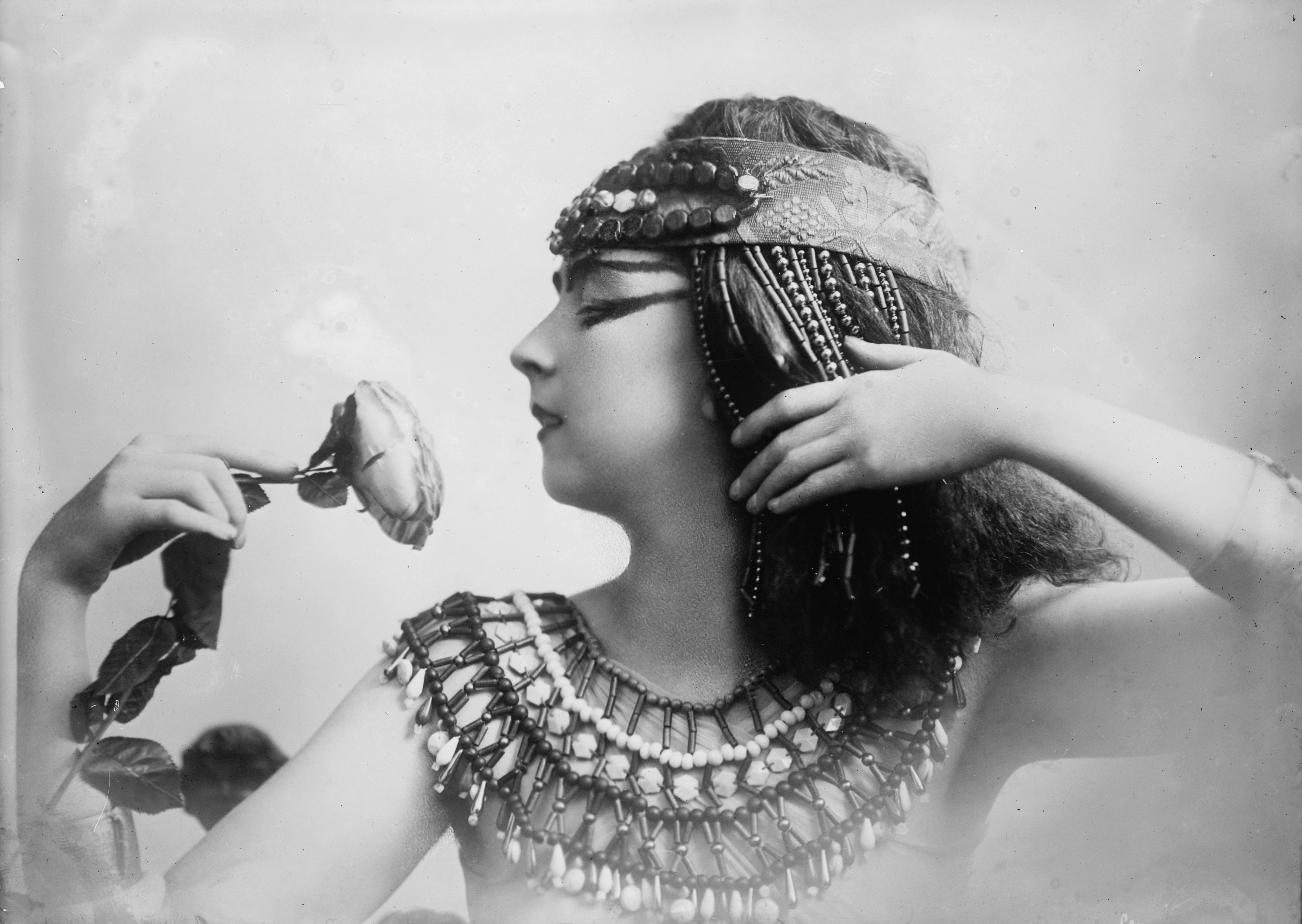 Ruth St. Denis holds a rose while wearing an Egyptian-influenced theatrical headdress and costume.