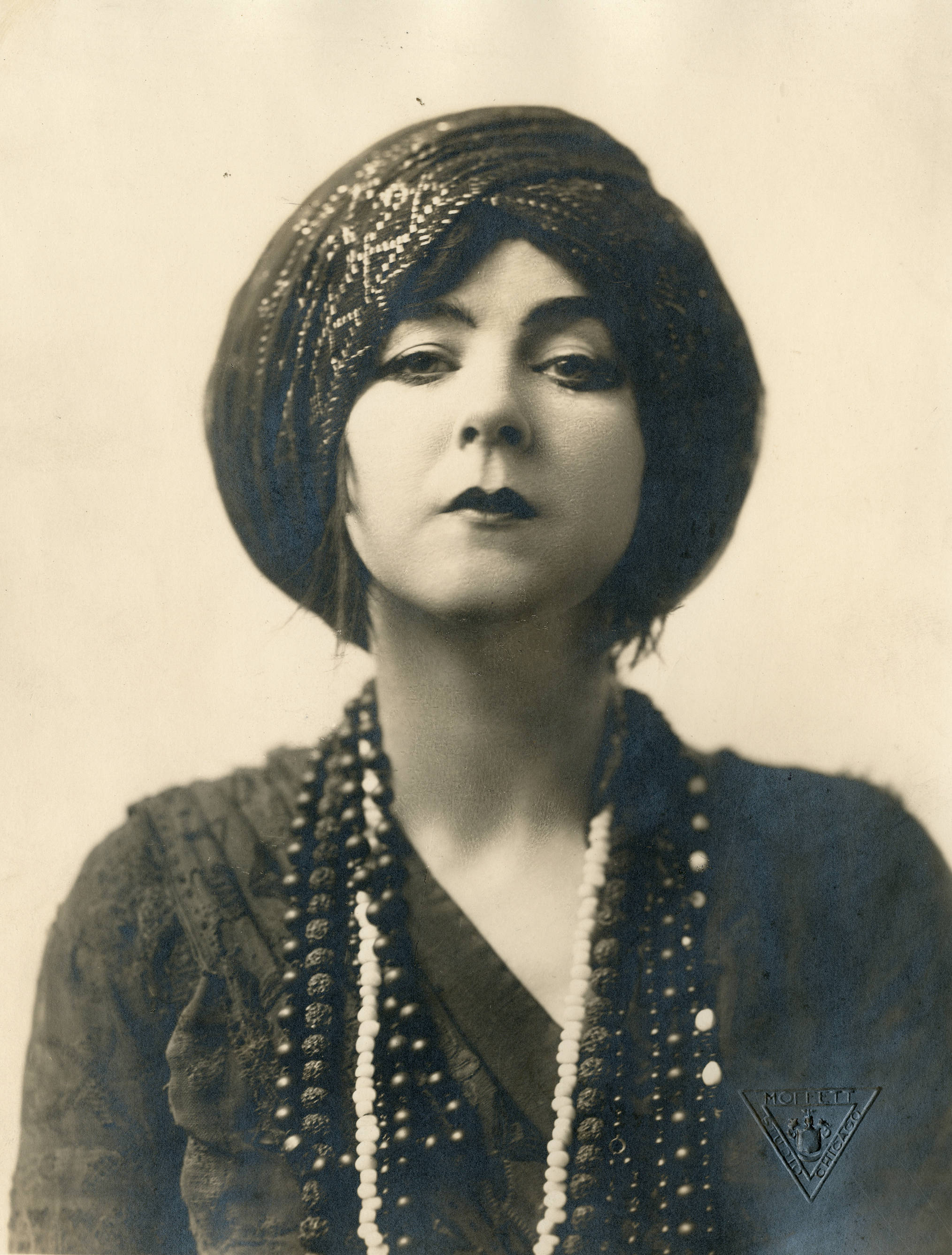 Close portrait of Ruth St. Denis wearing a jeweled head covering and layered necklaces in 1915.